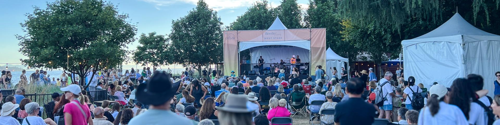 This is a wide-angle shot of an outdoor concert or festival on a clear, bright day. The scene is dominated by a large crowd of people seated on folding chairs, facing a performance stage. The stage, situated in the middle ground, is a temporary structure with a light-colored canopy and a banner that reads "Beedie Living WEST STAGE" and also displays "Arts". A band is performing on the stage, with several musicians visible.