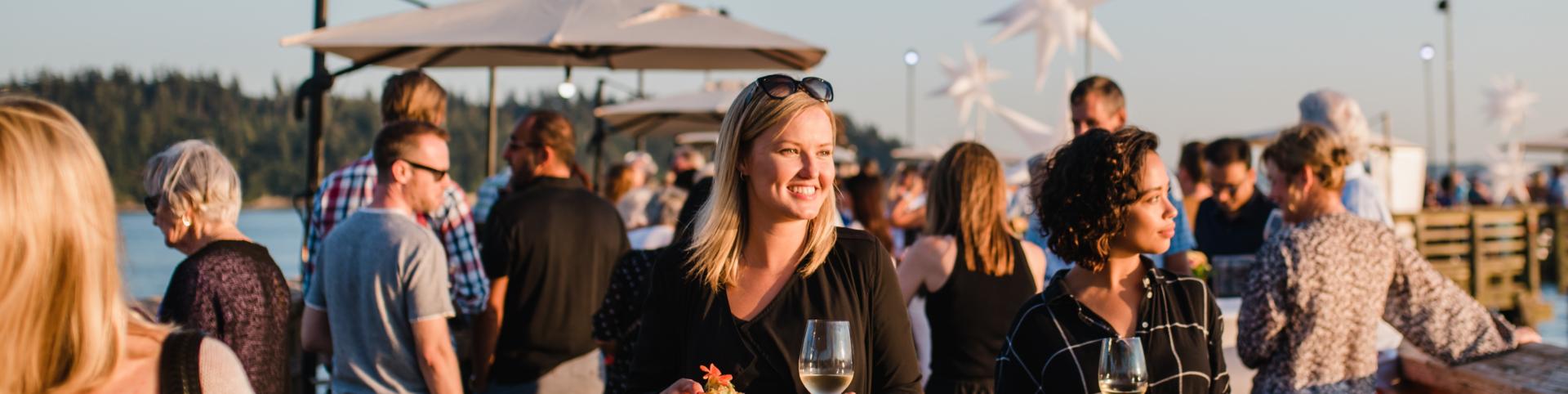 The image captures an outdoor social gathering at sunset, likely on a pier or boardwalk. In the foreground, two women are the focal points. The woman on the left, with blonde hair and sunglasses perched on her head, is smiling and looking to her right. She is holding a small white plate with a bit of food and a glass of white wine. She wears a black dress. Next to her, a woman with dark, curly hair is looking to her left, also holding a glass of white wine and a plate of food. She is wearing a black and whi