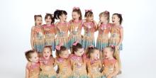 A group of young girls wearing colorful sequinned dresses, smiling and posing together for a photo.