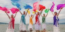 A group of dancers in colorful dresses and fans stands on rocks with the ocean in the background.