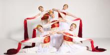 Five young women are positioned in a visually dynamic arrangement against a plain white background, suggesting a studio setting for a performance or photoshoot. They are all dressed in elaborate, traditional-inspired costumes featuring a white base with bright red bodices adorned with intricate gold embroidery and delicate gold fringe.