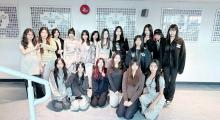 This image captures a group of approximately fifteen young women posing for a photo in what appears to be a school hallway. The group is arranged in two rows: a front row of seven women kneeling on the floor and a back row of eight women standing. They are all looking toward the camera, with various playful or neutral expressions. Several individuals are making hand gestures, such as peace signs or heart shapes, adding a casual and cheerful vibe to the photo.