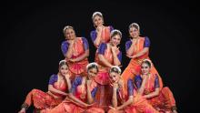 The image displays a group of eight young women performing what appears to be a traditional Indian dance. They are all dressed in vibrant, ornate costumes. Each dancer wears a blue, intricately patterned blouse with short sleeves and a full, orange skirt that has pink accents and pleats. Their attire is further adorned with heavy gold jewelry, including necklaces, earrings, bangles, and waistbands. They also wear decorative hairpieces, often with white elements. All the women have their hair styled up and a