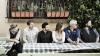 The image displays a group of six individuals, three men and three women, seated in a row at an outdoor table, likely a cafe or restaurant patio. They are all looking to their right, creating a unified gaze that draws the viewer's attention in that direction. The scene is set against a neutral beige wall and a window with decorative bars above it. Two terra-cotta planters filled with greenery and a few red flowers are positioned on a ledge beneath the window. To the right of the window, a dense wall of lush
