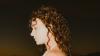 The image is a close-up, profile view of a young woman with voluminous, dark curly hair. Her head is tilted downwards, and her eyes are closed, suggesting a moment of reflection or peace. Her skin tone is fair, and she appears to be wearing subtle makeup, with a hint of eyeshadow. A delicate silver necklace is visible around her neck, and the top of a white garment, possibly a tank top or dress, can be seen.