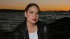 The image is a medium shot portrait of a young woman seated outdoors by the water, likely at dusk or dawn given the warm tones of the sky. The woman is positioned slightly off-center to the right, with her gaze directed straight at the camera. She has her hair pulled back neatly, revealing her face and ears. She is wearing a black leather jacket over a white t-shirt. Her makeup is noticeable, with emphasis on her eyes, which are adorned with false eyelashes, and her lips, painted in a bold reddish-orange hu