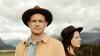 The image features a medium shot of two people, a man and a woman, positioned in the foreground against a blurred outdoor landscape.  The man is on the left side of the frame, facing the camera directly. He is bald and has a serious expression. He is wearing a wide-brimmed brown hat, a light blue denim shirt, and an open, mustard-yellow jacket. His jacket has a soft, textured appearance.
