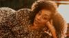 The image is a close-up, portrait-style photograph of a Black woman with curly dark brown hair, looking towards the camera. She is lying down, with her head resting on her left hand. The woman is wearing a shimmering dress covered in small, iridescent sequins that appear to be shades of bronze, gold, and copper, creating a textured and eye-catching garment. The sequins reflect the warm, golden light, adding to the overall glow of the image