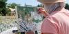 In a close-up, over-the-shoulder shot, an elderly person with white hair is painting outdoors. They are wearing a straw hat that casts a slight shadow over their face, and a salmon-pink t-shirt. The artist holds a thin paintbrush in their right hand, their fingers stained with paint, and is actively working on a canvas set on a dark easel. 